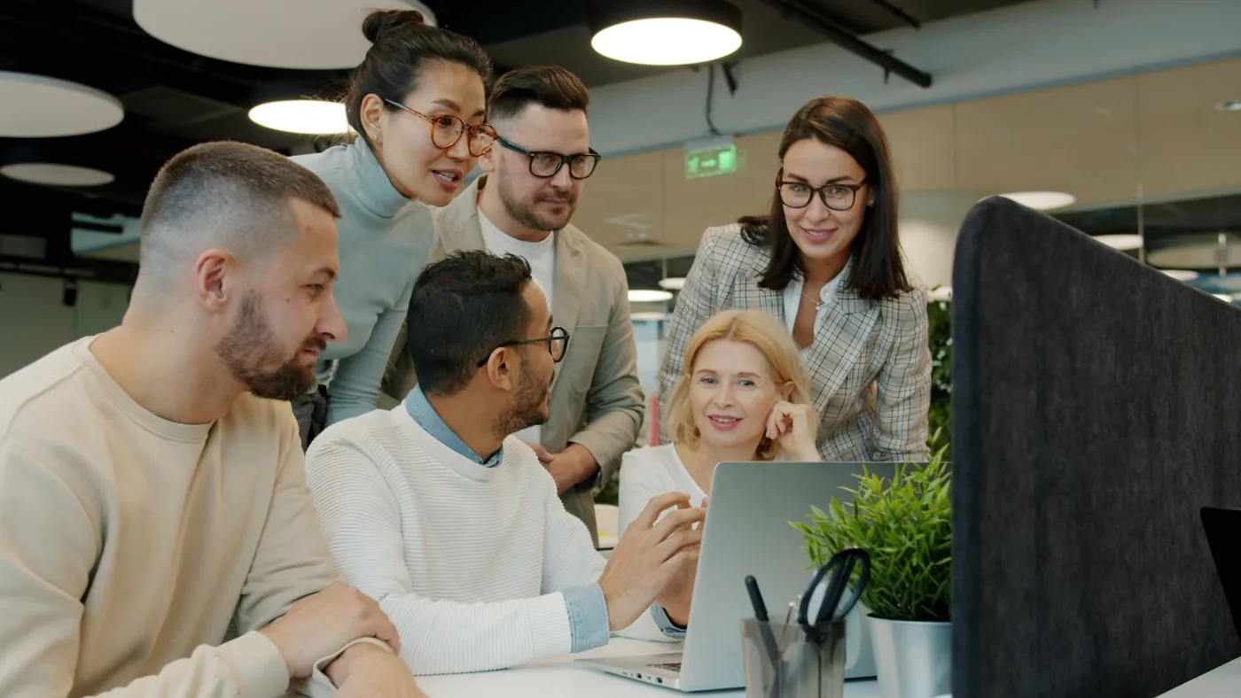 A diverse team of six professionals gathered around a laptop in a bright, modern office